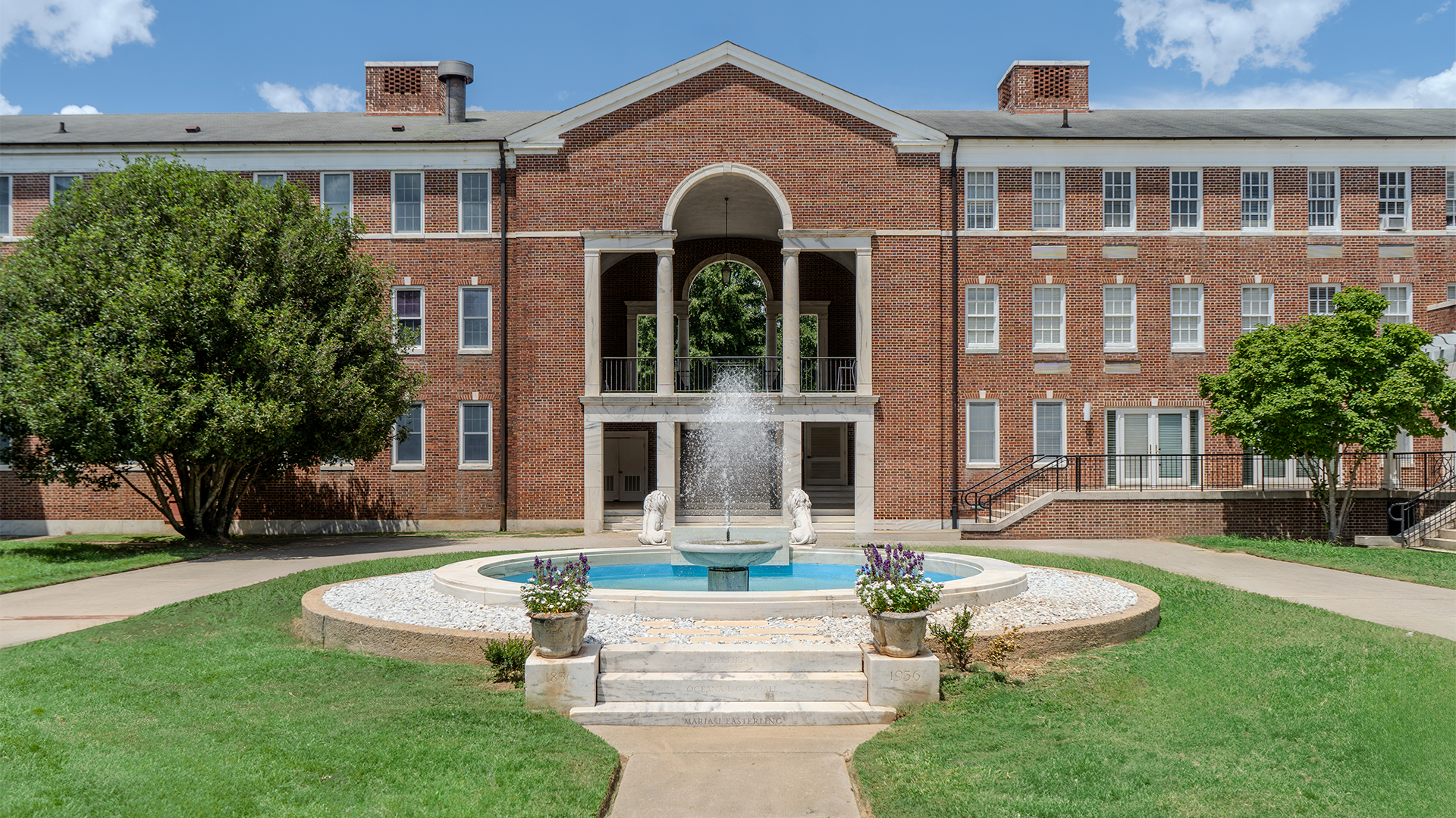 Campus Fountain Large brick campus building with an arched central entrance and a fountain in the foreground, surrounded by green lawns, trees, and flower planters under a bright blue sky.
