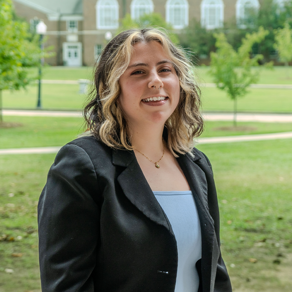 Mackie Headshot - Square Smiling young woman with short wavy hair featuring blonde highlights, standing outdoors on a campus lawn. She is wearing a black blazer over a light-colored top, with academic buildings and trees softly blurred in the background.