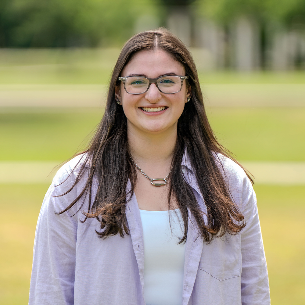 Skylar Headshot - Square Smiling young woman with long dark hair and glasses standing outdoors on a grassy campus lawn. She is wearing a light button-up shirt over a white top and a silver chain necklace, with trees softly blurred in the background.