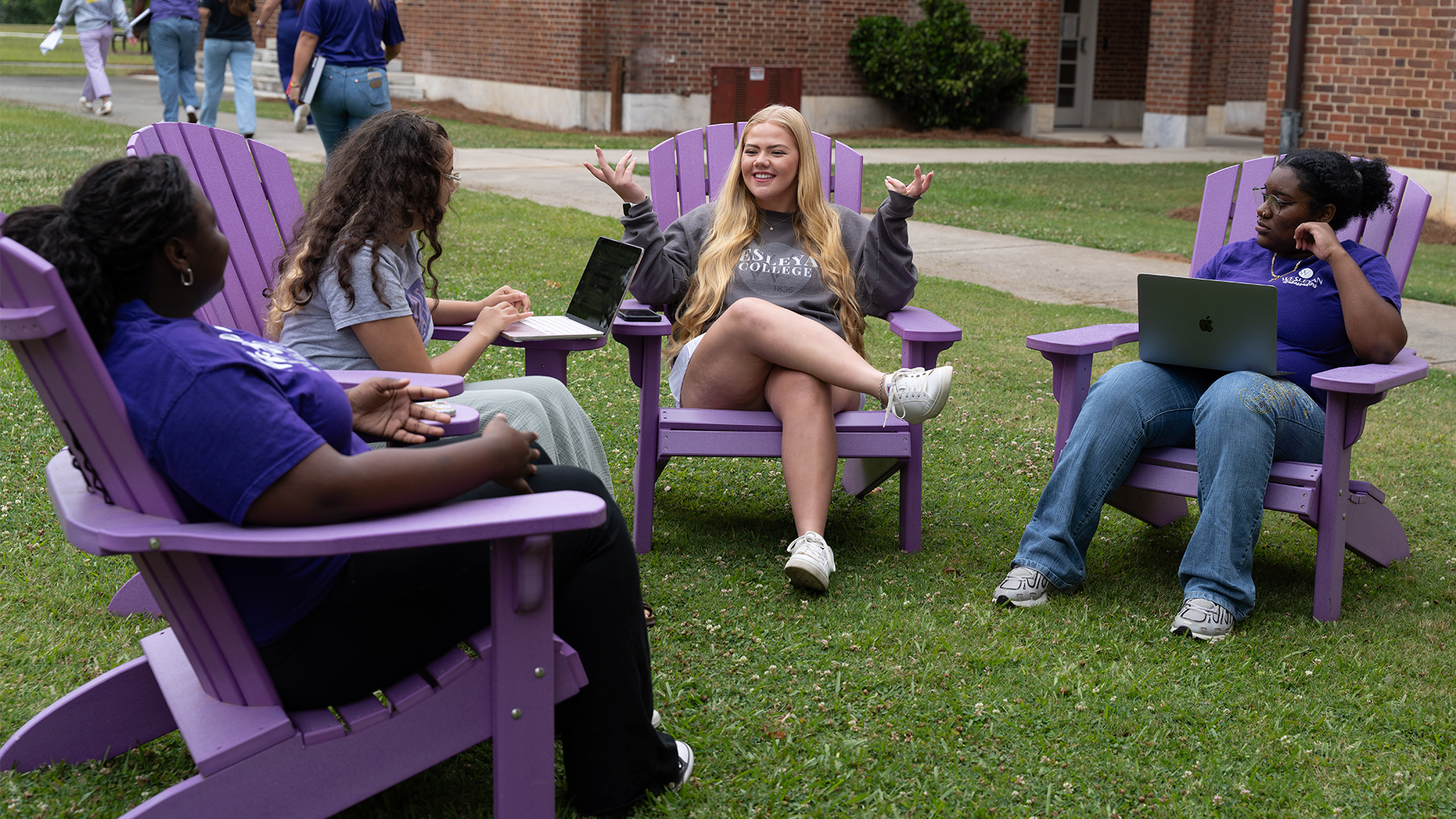 Students on Chairs Group of students sitting in purple Adirondack chairs on a campus lawn, talking and using laptops outside a brick academic building. One student gestures while speaking as the others listen and interact in a relaxed outdoor study setting.