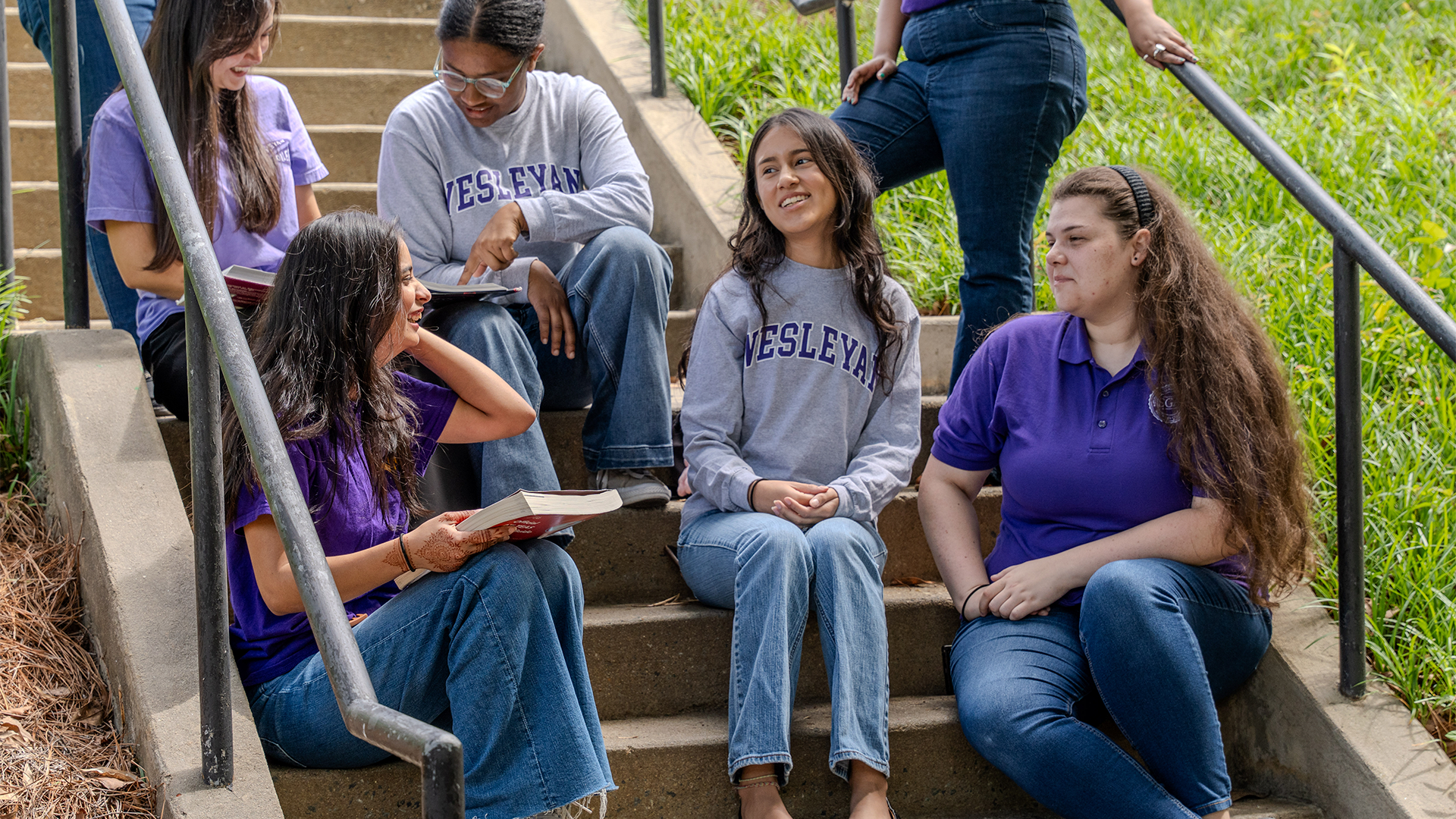 Students on Steps Group of students sitting together on outdoor campus steps, talking, reading, and smiling. Several wear Wesleyan shirts in purple and gray, creating a casual and collaborative study atmosphere surrounded by greenery.