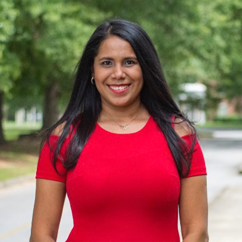 Yasmin Headshot - Square Smiling woman with long dark hair wearing a bright red dress, standing outdoors on a tree-lined campus walkway. The background is softly blurred with greenery and a road visible behind her.
